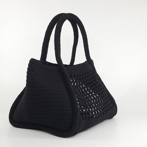 Op de afbeelding: Een zwarte gehaakte tote bag met een gaasontwerp aan de voorkant. De tas heeft twee handvatten.