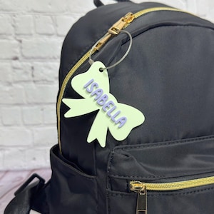 May include: A black backpack with gold zippers and a light green bow-shaped tag. The tag has the name "ISABELLA" in light purple. The backpack has a front pocket and adjustable straps.