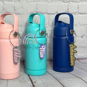 May include: Three insulated water bottles in pink, teal, and navy blue, each with a matching handle and a personalized name tag. The tags read "Chloe", "Mia", and "Sebastian". The bottles are designed for cold beverages.