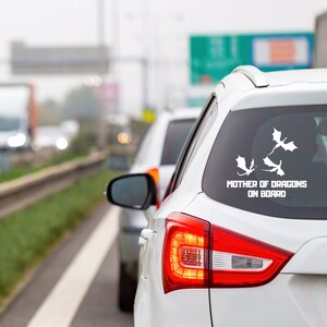 Pode incluir: Um carro branco com um adesivo na janela traseira que diz "Mother of Dragons On Board". O adesivo apresenta três silhuetas de dragões brancos.