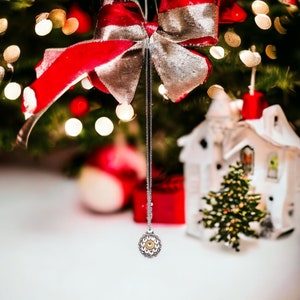 May include: A silver necklace with a round pendant featuring a gold center and a decorative design hangs from a red and silver bow on a Christmas tree.