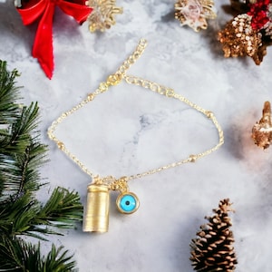 May include: A gold chain bracelet with a small gold cylinder charm and a blue evil eye charm. The bracelet is on a white marble surface.