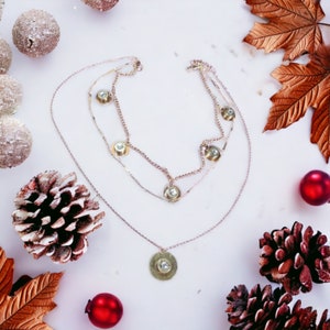 May include: A layered silver necklace with a gold pendant featuring a circular design with a small crystal in the center. The necklace is surrounded by pine cones and red ornaments.