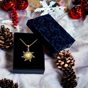 May include: A gold sun pendant necklace with a white stone in the center, displayed in a black velvet jewelry box. The box is open and the necklace is lying inside. The box is surrounded by pine cones and red ornaments.