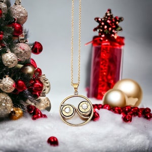 May include: A gold necklace with a pendant featuring two bullet casings with rhinestones. The necklace is displayed against a background of a Christmas tree with red and gold ornaments and a red and gold gift box.