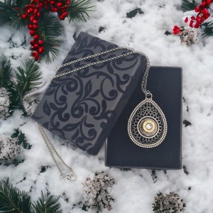 May include: A silver necklace with a teardrop-shaped pendant featuring a bullet casing and a clear rhinestone. The necklace is displayed in a black velvet box with a floral pattern.