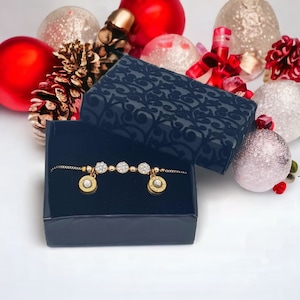 May include: A gold bracelet with small round charms set with clear rhinestones. The bracelet is displayed in a black velvet box with a decorative pattern. The box is surrounded by red and white Christmas ornaments.