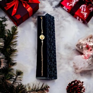 May include: A gold chain bracelet with a round pendant featuring a floral design and a single clear stone in the center. The bracelet is displayed in a black velvet box with a floral pattern.