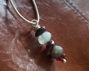 Op de afbeelding: Een zilveren ketting met een hanger gemaakt van gelaagde edelstenen. De hanger heeft een lichtblauwe steen, een donkergrijze steen, een donkerrode steen en een kleinere donkerrode steen.