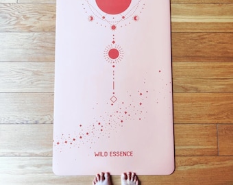 Peut inclure: Un tapis de yoga rose avec un motif de soleil et de lune rouge et le texte "WILD ESSENCE".