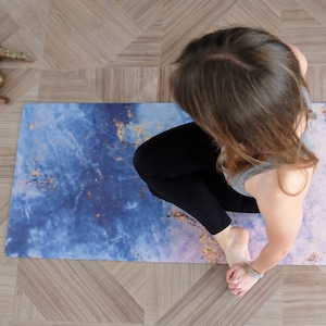 Op de afbeelding: Een persoon zit in een yogapose op een blauwe en paarse yogamat met gouden vlokken. De mat heeft een marmerachtig patroon.