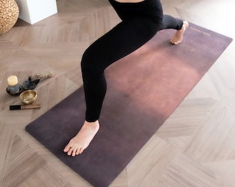 Peut inclure: Un tapis de yoga avec un dégradé de couleurs, passant du violet foncé au rose clair. Le tapis est rectangulaire et porte l'inscription "WILD SECRETS" sur un bord. Une personne pratique le yoga sur le tapis.