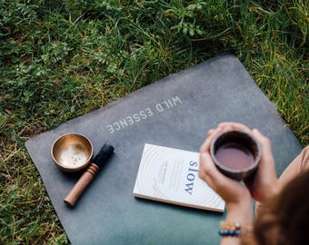 May include: A dark green rectangular yoga mat with the text WILD ESSENCE printed in white. On the mat sits a small metallic gold singing bowl with a wooden mallet and a white book titled slow by Lee Holden. A dark ceramic mug is also present.