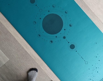 Peut inclure: Tapis de yoga turquoise avec un motif céleste de soleils, de lunes et d'étoiles. Le tapis porte l'inscription "WILD ESS". Le tapis est posé sur un sol en bois clair. La personne porte un pantalon gris et des chaussettes rayées.