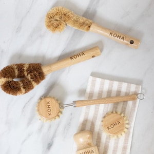 May include: Set of four wooden cleaning brushes with natural bristles. Each brush has a wooden handle with the word "KOHA" engraved on it. The brushes are arranged on a white marble surface and a striped fabric.