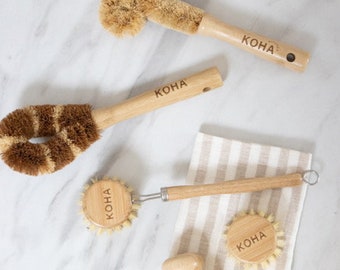 May include: Set of four wooden cleaning brushes with natural bristles. Each brush has a wooden handle with the word "KOHA" engraved on it. The brushes are arranged on a white marble surface and a striped fabric.