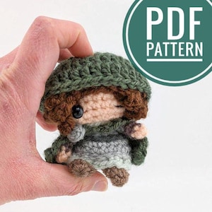 Può includere: Bambola amigurumi all'uncinetto con capelli castani, un mantello con cappuccio verde e una tunica grigia. La bambola tiene un piccolo oggetto in mano.