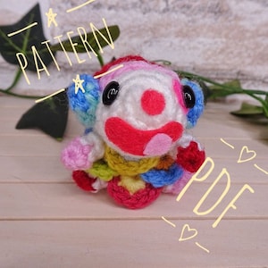 Puede incluir: Un muñeco de payaso de crochet colorido con una nariz roja y una gran sonrisa. El muñeco lleva un sombrero y una bufanda multicolores. La imagen incluye el texto "PATTERN - PDF - ".