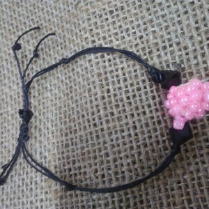Könnte beinhalten: Ein schwarzes Armband aus Schnur mit einem rosa Perlenanhänger. Das Armband hat einen einfachen Knotenverschluss.