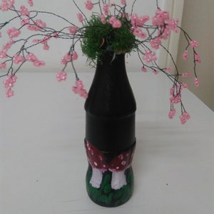 May include: A black glass bottle with a green moss top and pink beaded flowers. The bottle is painted with red and white mushrooms and green grass.