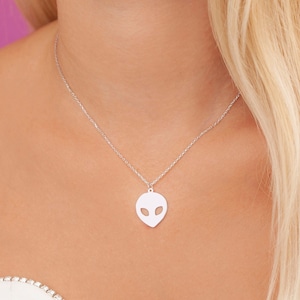 May include: A silver-colored necklace featuring a pendant shaped like an alien head with large oval eyes. The pendant hangs from a delicate chain. The background is a soft pink.