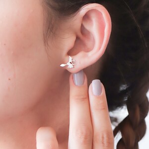May include: Silver witch on a broomstick earrings. The earrings are shown on a person's ear with a finger pointing to the earring.