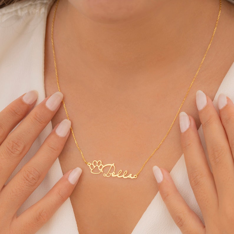 May include: Gold necklace with a paw print charm and the name "Bella" written in cursive.