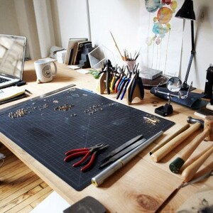 May include: A workspace with a black cutting mat, various jewelry making tools, and a white mug. The tools include pliers, a magnifying glass, a hammer, and a file. There are also small metal rings and beads scattered on the mat.