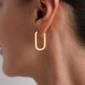 May include: A close-up of a gold-colored, rectangular hoop earring. The earring is a simple, modern design with a slight curve and a small gap at the bottom. The earring is worn on the ear.