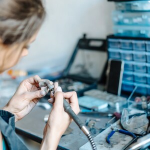 May include: A person is working on a piece of jewelry with a small tool. They are holding the jewelry in their left hand and using the tool in their right hand. The jewelry is silver and has a small, intricate design.