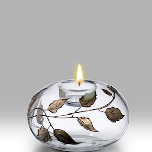 Op de afbeelding: Transparante glazen kandelaar met een brandende waxinelichtje. De houder heeft een ovale vorm en is versierd met een goudkleurig blad- en rankenontwerp. De kaarsvlam schijnt tegen een neutrale achtergrond.