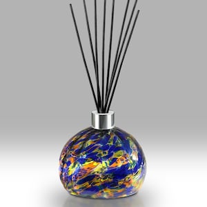 May include: A decorative glass diffuser with a round, colorful base. The base features a blend of blue, yellow, and orange hues. Black reeds extend upwards from a silver-colored collar. The diffuser is designed for home fragrance.