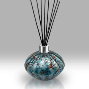 Puede incluir: Un difusor de caña decorativo con una botella de vidrio redonda con un patrón azul y rojo. La botella tiene un collar de metal plateado y está rematada con varios palitos difusores negros. El difusor está diseñado para la fragancia del hogar.