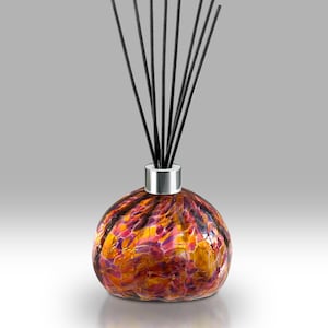 May include: A decorative glass diffuser with a round, speckled body in shades of orange, yellow, and purple. The diffuser has a silver collar and is topped with several black reeds, designed for releasing fragrance.