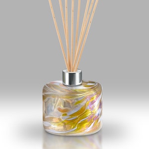 Op de afbeelding: Een glazen diffuserfles met een swirl-patroon in tinten goud, wit en roze. De fles heeft een zilveren dop en is gevuld met een heldere vloeistof. Houten diffuserstokjes zijn in de fles gestoken.