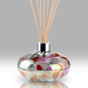 Op de afbeelding: Een ronde glazen diffuserfles met een zilveren dop en een kleurrijk abstract ontwerp. De fles is gevuld met een heldere vloeistof en heeft houten stokjes die in de bovenkant zijn gestoken.