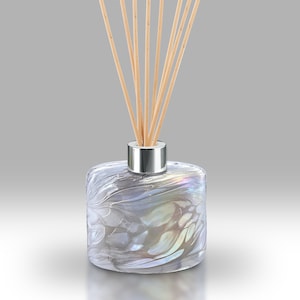 May include: A pearlescent glass reed diffuser with a silver-coloured collar. The bottle, filled with liquid, has a square shape with rounded edges. Light brown reeds are inserted into the bottle. The diffuser is designed for home fragrance.