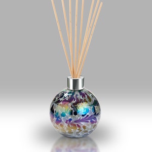 Op de afbeelding: Een ronde glazen diffuserfles met een zilveren dop en iriserende blauwe, paarse en gouden swirls. De fles is gevuld met een heldere vloeistof en heeft houten stokjes die uit de bovenkant steken.