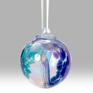 Puede incluir: Una bola de Navidad de cristal transparente con una cinta blanca. El adorno redondo presenta una mezcla de colores azules, turquesas y morados. El cristal tiene una cualidad iridiscente, reflejando la luz. El adorno est&aacute; dise&ntilde;ado para colgar.