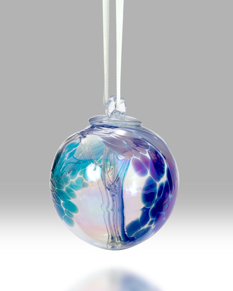 Puede incluir: Una bola de Navidad de cristal transparente con una cinta blanca. El adorno redondo presenta una mezcla de colores azules, turquesas y morados. El cristal tiene una cualidad iridiscente, reflejando la luz. El adorno est&aacute; dise&ntilde;ado para colgar.