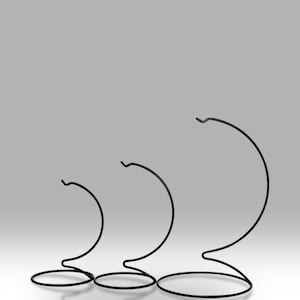 Puede incluir: Tres soportes de exhibición de metal negro de diferentes alturas. Cada soporte tiene una base circular, un cuerpo curvo y un gancho en la parte superior. Los soportes están dispuestos en fila, el más alto a la derecha.