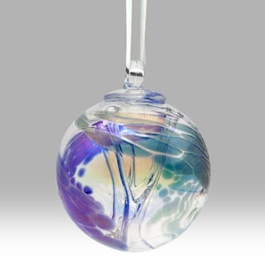 Puede incluir: Un adorno de vidrio transparente con remolinos de colores azul, morado y verde. El adorno tiene un lazo en la parte superior con una cinta blanca para colgar. El vidrio tiene una cualidad iridiscente.