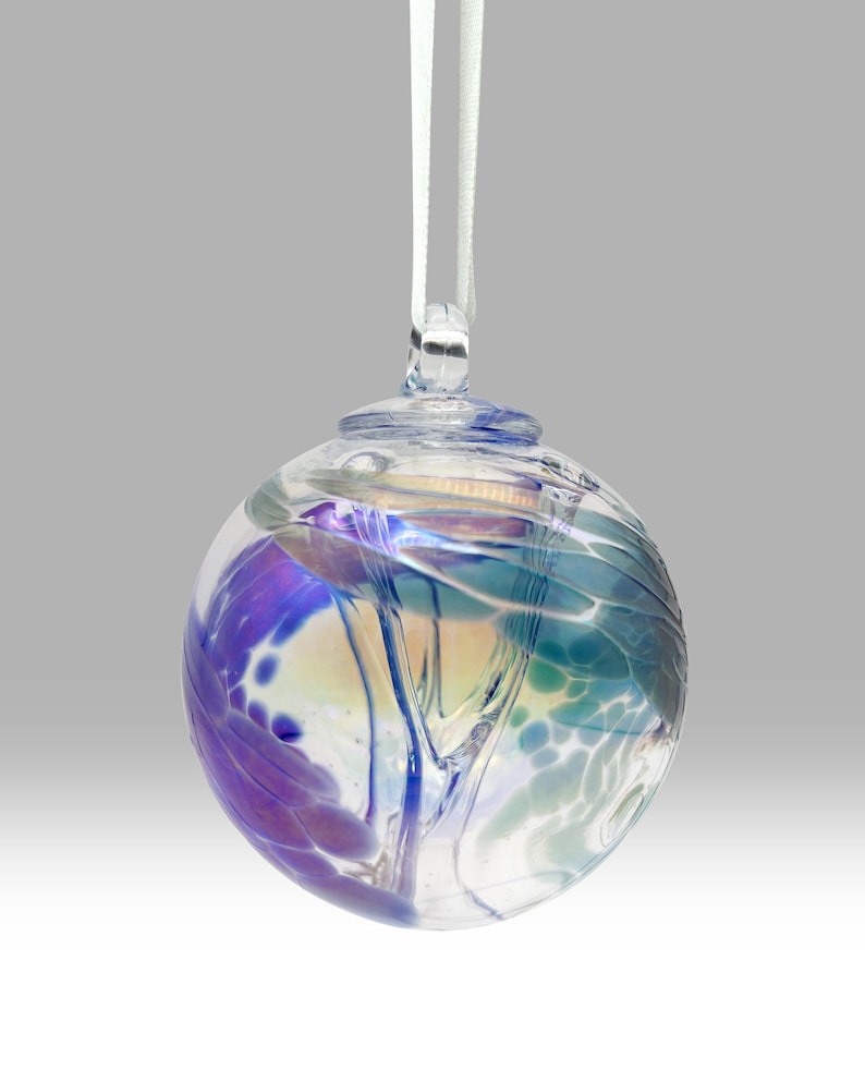 Puede incluir: Un adorno de vidrio transparente con remolinos de colores azul, morado y verde. El adorno tiene un lazo en la parte superior con una cinta blanca para colgar. El vidrio tiene una cualidad iridiscente.