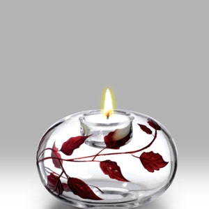 May include: A clear glass candle holder with a red leaf design. The holder has a small, round opening for a tea light candle.