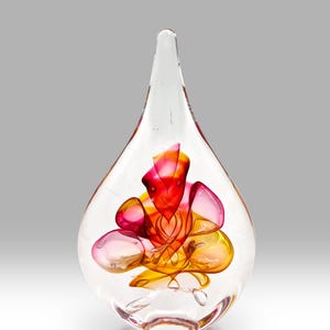 Può includere: Un vaso di vetro trasparente a forma di lacrima con un fiore di vetro rosa e arancione all'interno.