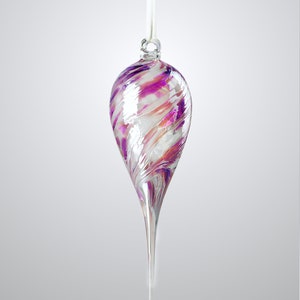 Puede incluir: Adorno de vidrio en forma de lágrima con una cinta blanca para colgar. El adorno presenta patrones arremolinados de blanco, rosa y morado. La parte inferior se estrecha hasta una punta larga y puntiaguda. Un artículo decorativo para las fiestas.