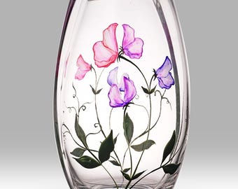 May include: Clear glass vase with a hand-painted floral design. The vase features pink and purple flowers with green leaves and stems. The vase is oval-shaped and sits on a white surface.