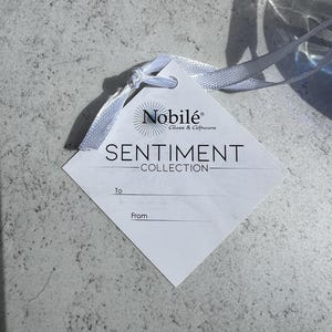 Puede incluir: Una etiqueta blanca en forma de diamante con el texto "SENTIMENT COLLECTION" y la marca "Nobil&eacute; Glass & Giftware". La etiqueta est&aacute; atada con una cinta blanca. El fondo es una superficie texturizada de color gris claro.