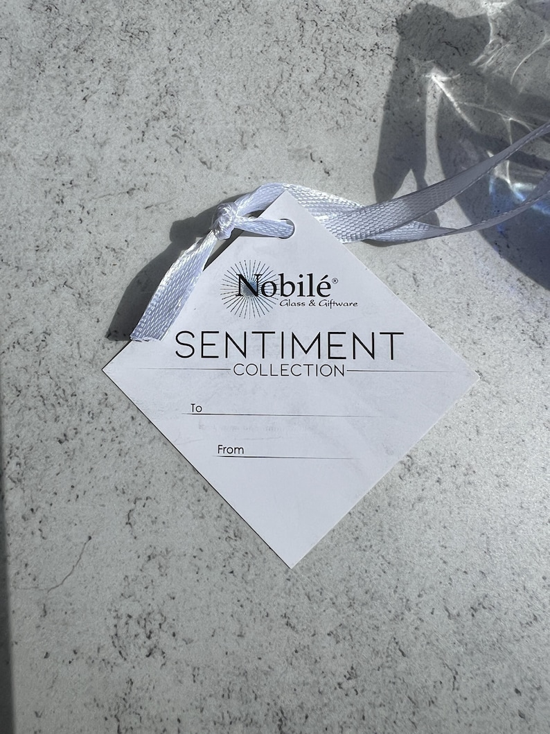 Puede incluir: Una etiqueta blanca en forma de diamante con el texto "SENTIMENT COLLECTION" y la marca "Nobil&eacute; Glass & Giftware". La etiqueta est&aacute; atada con una cinta blanca. El fondo es una superficie texturizada de color gris claro.