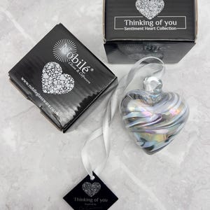 Op de afbeelding: Een hartvormig glazen ornament met iriserende kleuren, opgehangen aan een wit lint. Twee zwarte dozen met witte hartontwerpen en de tekst "Thinking of you" zijn zichtbaar. Een zwart label met dezelfde tekst is ook aanwezig.